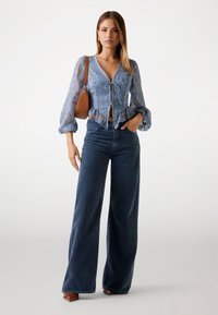 Lichtblauw geborduurde crop top met pofmouwen, met een geknoopt voorpand; gecombineerd met hoge, donkere blauwe wijde jeans en een bruine handtas.
