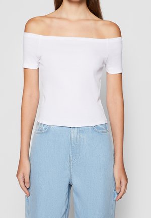 Une personne portant un haut blanc à manches courtes et épaules dénudées ainsi qu'un jean taille haute bleu clair, sur un fond gris uni.