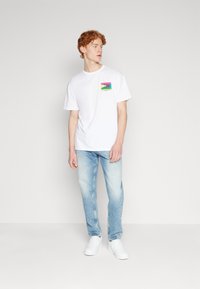 Camiseta de algodón blanca de manga corta, con un parche gráfico colorido. Combinada con jeans de mezclilla azul claro y zapatillas blancas.