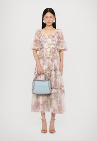 Vestito in chiffon floreale di un delicato viola pastello con maniche a volant; borsa blu abbinata con dettagli in pelle; modello che indossa sandali nudi con cinturini.