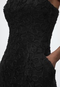 Κοντινή λήψη μαύρου φόρεματος με υφή και ανάγλυφη κεντητική λουλουδιών, με ένα χέρι τοποθετημένο σε πλαϊνή τσέπη.