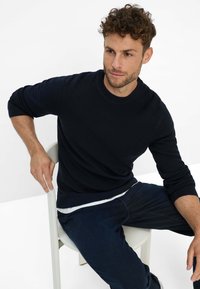Marineblauwe gebreide trui met ronde hals en geribbelde textuur, gedragen over een witte t-shirt, gecombineerd met donkere jeans.