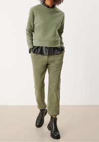 Felpa verde oliva, indossata sopra una blusa in pelle nera, abbinata a pantaloni verde oliva e stivaletti neri. Design semplice e moderno.