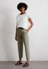 Witte katoenen T-shirt met ronde halslijn, licht olijfgroene tapered broek en zwarte sandalen met een gouden gesp. Eenvoudig ontwerp en kleuren.
