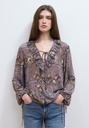 Donna con capelli lunghi castani indossa una camicetta con motivi decorati con volant e lacci, abbinata a jeans blu, in piedi davanti a uno sfondo chiaro e semplice.