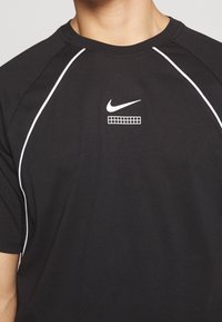 Musta Nike t-paita, jossa valkoisia yksityiskohtia, lyhyet hihat ja pyöreä pääntie. Rintakehällä on logo ja graafinen kuvio.