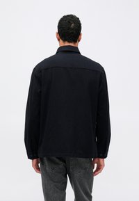 Schwarz langärmliges Hemd mit abgerundetem Saum und geknöpften Manschetten, hergestellt aus einem weichen Stoff. Mit Kragen und Rückenpasse ausgestattet.