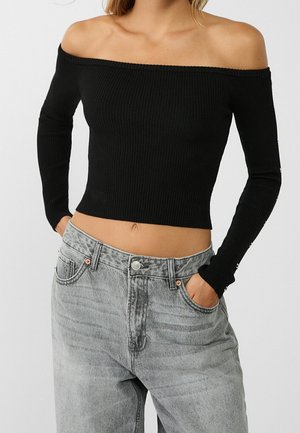 Vrouw die een zwart geribd off-shoulder crop top met lange mouwen en een hoge taille lichtgrijze gewassen spijkerbroek draagt, met haar handen in haar zakken.