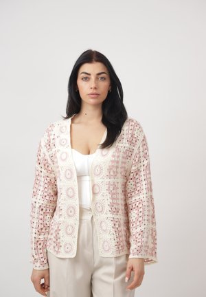 Vrouw met lang donker haar, gekleed in een witte top en beige broek, gecombineerd met een roze-witte gehaakte open-front cardigan.