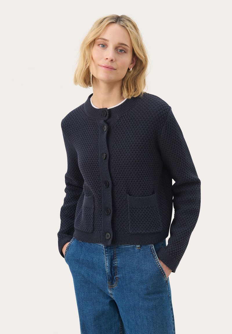 Merihenkinen neulottu cardigan, jossa on teksturoitu kuosi, pyöreä kaula-aukko, keskellä napit ja kaksi etutaskua. Yhdistettyinä sinisiin farkkuihin.