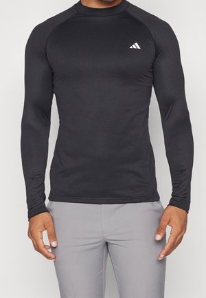 Mies, der ein enganliegendes schwarzes langärmeliges Sportshirt mit einem kleinen weißen Adidas-Logo auf der Brust und hellgrauen Hosen trägt.