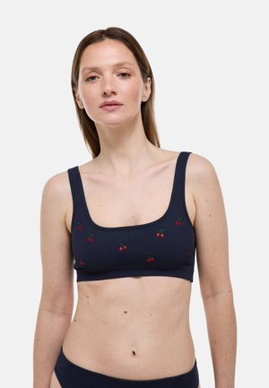 Femme portant un crop top côtelé bleu marine avec de petits motifs de cerises rouges sur un fond blanc uni.