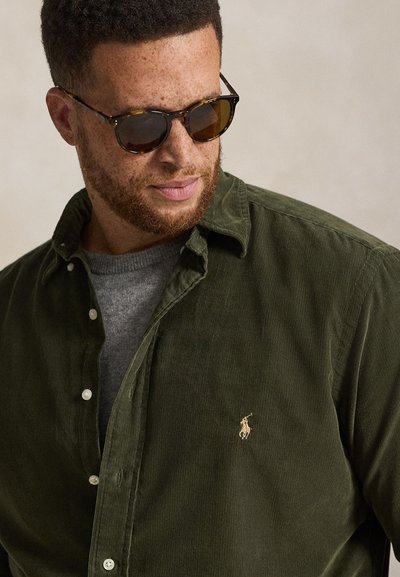 Homme aux cheveux courts et bouclés avec une barbe, portant des lunettes de soleil écaille de tortue, une chemise en velours côtelé verte sur un pull gris.