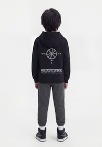 Παιδί με πλάτη φοράει μαύρο φούτερ Jordan με πυξίδα και λογότυπο, γκρι φόρμα, λευκές κάλτσες και μαύρα αθλητικά παπούτσια σε απλό φόντο.