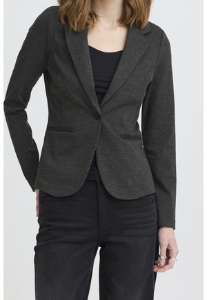 Frau trägt einen taillierten dunkelgrauen Blazer über einem schwarzen Top und schwarzen Hosen, steht mit einer Hand in der Tasche und der anderen entspannt.