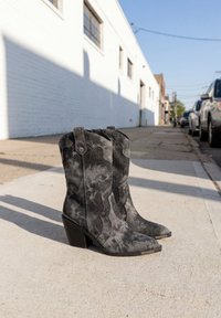 Paire de bottes de cowboy en suède noir avec talons blocs placées sur un trottoir urbain ensoleillé près de voitures garées et d'un bâtiment blanc.