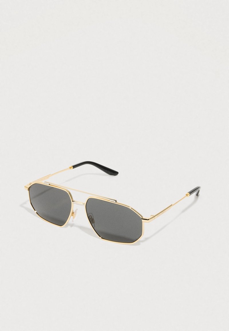 Óculos de sol de metal dourado com formato hexagonal, lentes cinza escuro e pontas das hastes em plástico preto. Apresenta um design de armação fina e plaquetas ajustáveis no nariz.