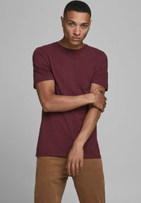 Bordeauxfarbenes T-Shirt mit kurzen Ärmeln aus einer weichen Baumwollmischung, taillierte Form, Rundhalsausschnitt. Das Modell zeigt die Arme verschränkt und trägt ein schlichtes goldenes Armband.