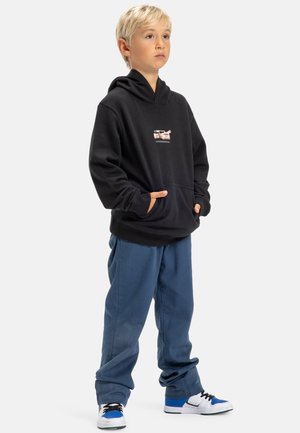 Schwarzer Hoodie mit Fronttasche und kleinem Grafikdruck, kombiniert mit blauen Hosen und weißen Sneakers mit blauen Akzenten. Weiche Stofftextur.
