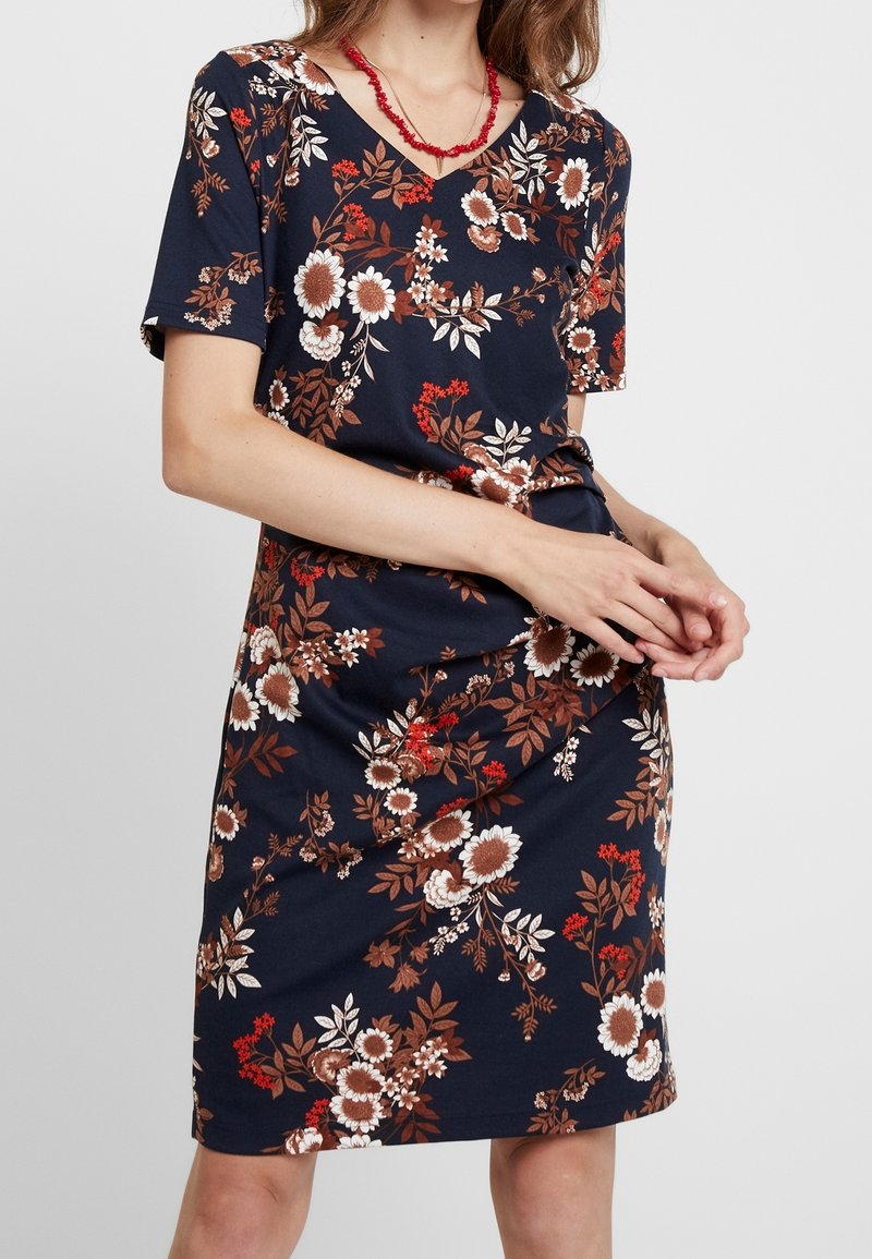 Femme portant une robe bleu marine mi-genoux avec des motifs floraux marron, blancs et rouges, et un collier de perles rouges, les mains jointes à la taille.