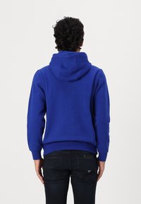 Blaue Kapuzenjacke aus einem Baumwollmischgewebe, mit großer Kapuze, gerippten Bündchen und Saum. Getragen mit dunklen Jeans, von hinten betrachtet.