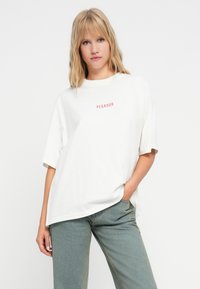 Λευκό φαρδύ μπλουζάκι με κοντά μανίκια, που φέρει κόκκινο κείμενο "PEGADOR" στο στήθος. Λείος ύφασμα και χαλαρή εφαρμογή. Φοριέται με πράσινο παντελόνι.