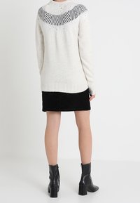 Stickad tröja i crème med svarta geometriska mönster vid oket, kombinerad med en svart kjol och blanka ankelstövlar, sedd bakifrån.