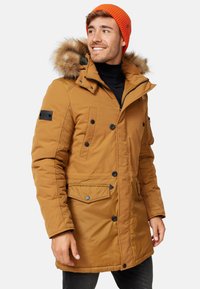 Parka isolée marron avec une capuche en fausse fourrure, plusieurs poches avant, boutons noirs et un bonnet en tricot orange.