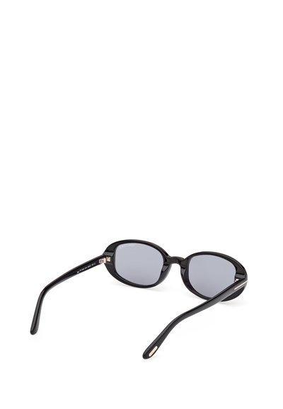 Occhiali da sole neri con montatura ovale e lenti grigie scure poggiati aperti su una superficie bianca, inclinati per mostrare l'interno e le aste.