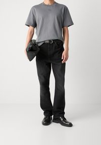 Grijze T-shirt, zwarte denim jeans en zwarte leren schoenen. De T-shirt heeft een ronde hals; de jeans hebben een rechte snit. Houdt een zwarte clutch bij zich.