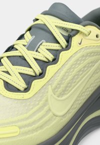 Αθλητικό παπούτσι Nike με ανοιχτό κίτρινο και γκρι χρώμα, υφασμένο συνθετικό επάνω μέρος, κυματιστά διακοσμητικά στοιχεία και επίπεδα κορδόνια.