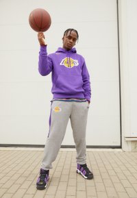 Felpa viola con logo "Lakers", pantaloni della tuta grigi con accenti blu e sneakers nere/viola. In mano una pallacanestro, su uno sfondo neutro.