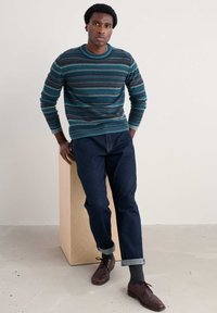 Randig stickad tröja i petrol, grått och brunt. Mörka denimjeans med uppvikta muddar. Modellen poserar mot en träpelare på en ljus bakgrund.