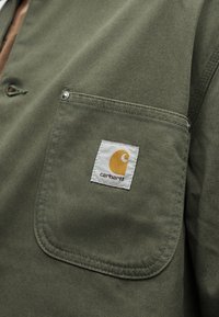 Giacca in tessuto verde con una tasca frontale decorata da una patch con il logo Carhartt bianco e giallo cucita sulla tasca.