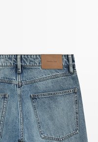 Vue arrière d'un jean en denim bleu avec deux poches et une étiquette rectangulaire marron cousue sur la ceinture.