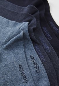 Štiri pare zloženih nogavic Calvin Klein v različnih odtenkih modre, urejenih drug ob drugem na beli površini.