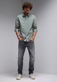 Vihreä raidallinen nappipaita, jossa on rintatasku, yhdistettynä kuluneisiin harmaisiin farkkuihin ja valkoisiin tennareihin. Malli seisoo kädet taskuissa.