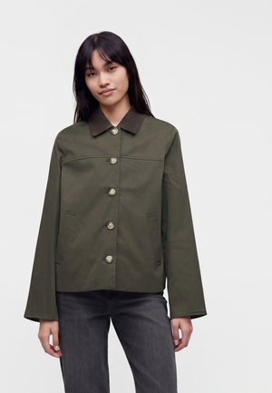 Frau mit glattem dunklem Haar, die eine olivgrüne Jacke mit großen Knöpfen und schwarze Jeans trägt, steht vor einem schlichten hellen Hintergrund.