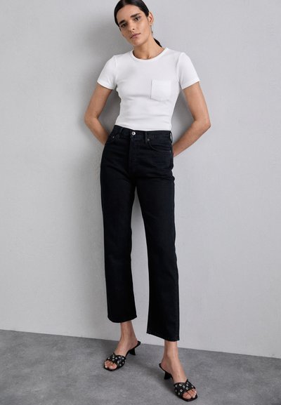 Black high-waisted jeans, straight leg, paired with a fitted white ribbed t-shirt featuring a chest pocket. Black heeled sandals with accents.