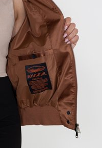 De binnenkant van een bruin bomberjack, met een soepele satijnen textuur, een ritsluiting en een patchlabel met oranje tekst en logo.