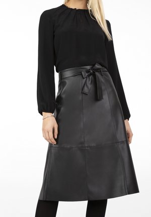 Kobieta w czarnej bluzce z długim rękawem i czarnej spódnicy z ekoskóry do kolan, z paskiem wiązanym na kokardę w talii.