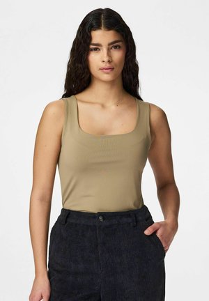 Kvinne med langt, bølgete mørkt hår iført en ermeløs beige topp og svarte kordfløyelsbukser, stående med den ene hånden i lommen.