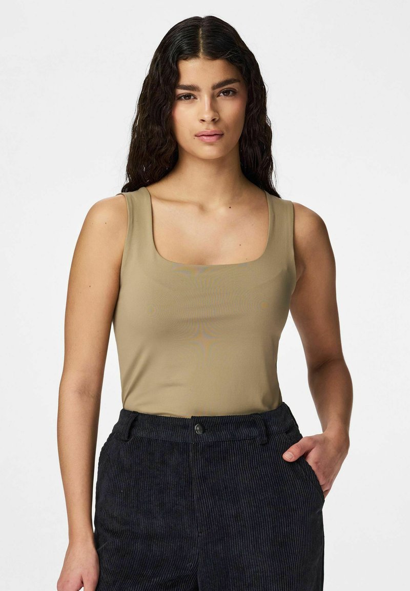 Femeie cu păr lung, ondulat și închis la culoare, purtând un top bej fără mâneci și pantaloni negri din catifea cu dungi, stând cu o mână în buzunar.