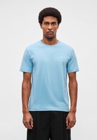 Világoskék pamut póló rövid ujjakkal, kerek nyakkivágással és egy kis fehér logóval a bal mellrészen. Egyszerű, dísztelen design.