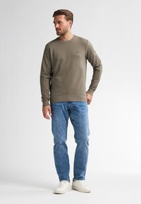 Felpa verde oliva con scollo rotondo, polsini e orlo a costine, abbinata a jeans azzurri chiari e sneakers bianche. Dettaglio del logo ricamato semplice.
