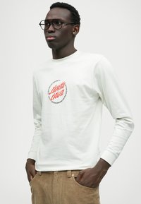 Long-sleeve white cotton T-shirt featuring a red and black graphic logo centered on the chest. Model wears brown corduroy pants.