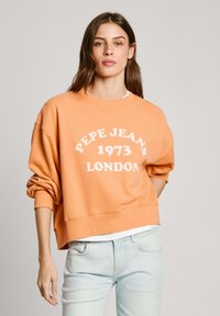 Oranžs pārāk liels džemperis ar baltu tekstu "PEPE JEAN'S 1973 LONDON". Ar noapaļotu apkakli un brīviem uzmetņu piedurkņu galiem. Gaiši zilas džinsas.