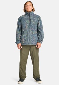 Fleece pullover in blue with geometric patterns, half-zip front, and ribbed cuffs. Paired with olive green pants and black sneakers.