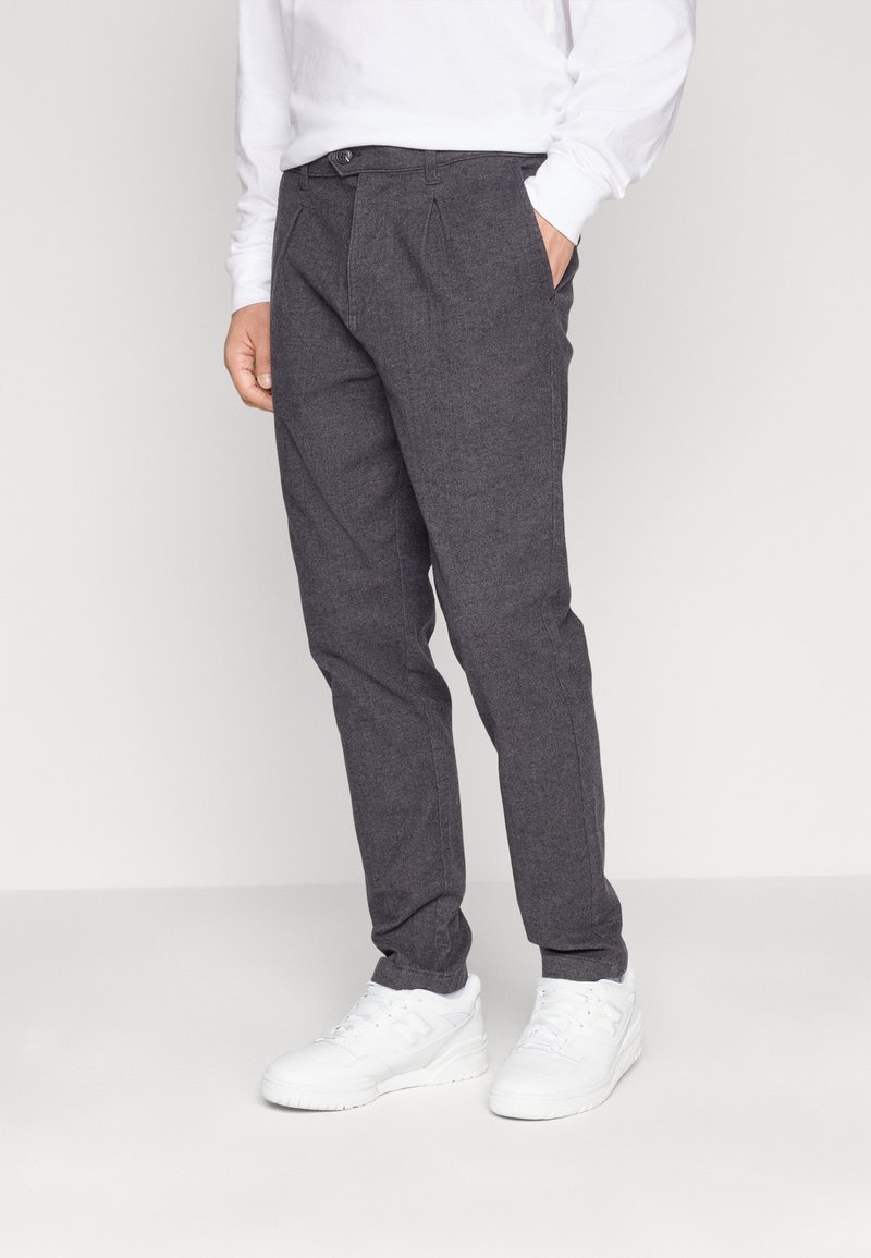 Pantaloni grigi realizzati in tessuto testurizzato, con una vestibilità su misura, caratterizzati da pieghe e tasche laterali, indossati con sneakers bianche.