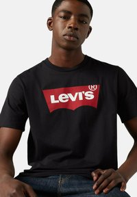 Joven vestido con una camiseta negra de Levi's con un logo rojo y blanco, sentado con una mano apoyada en su muslo frente a un fondo liso.
